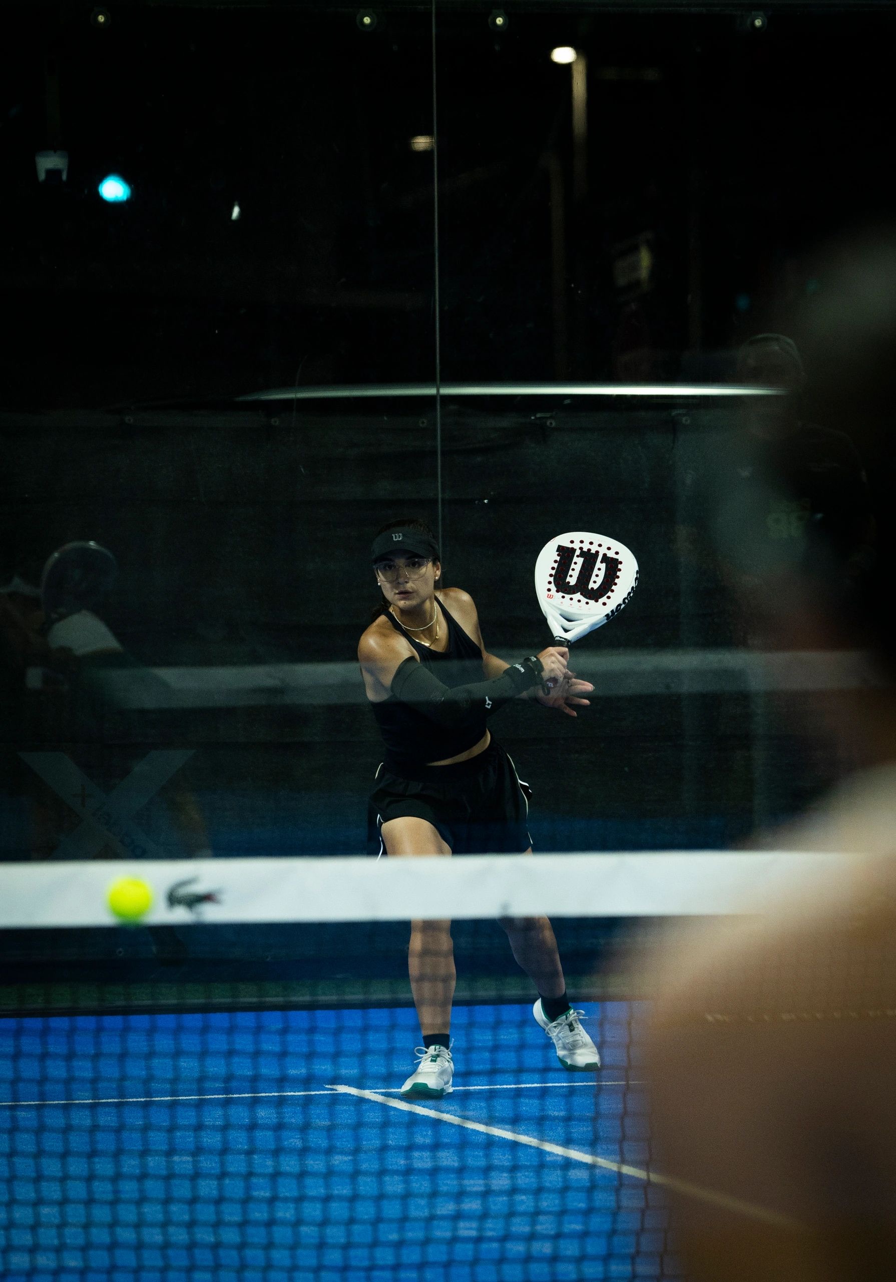 Ysa playing padel at night, seen through the glass wall of the court